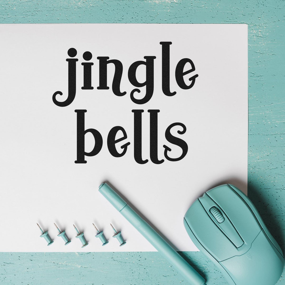 Mint green stamp and seal set with black jingle bells lettering, teal pen, and pins on white paper.
