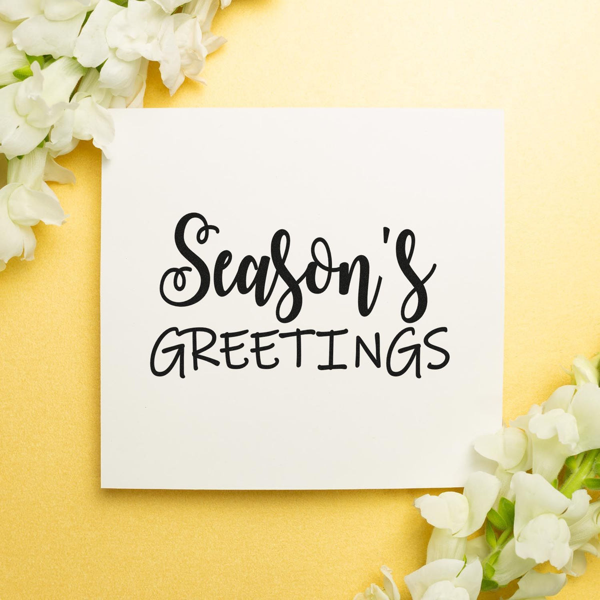 Seasons Greetings stamp and seal on white card, black lettering, framed by white flowers on a soft yellow background.