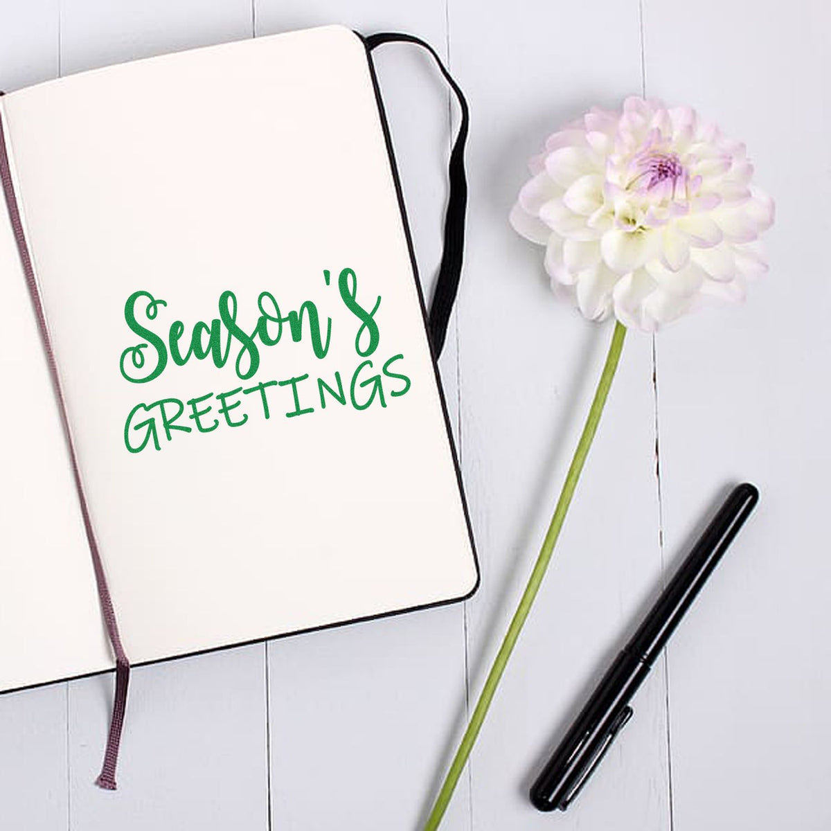 Green Seasons Greetings stamp and seal on cream notebook, with white flower and black pen.