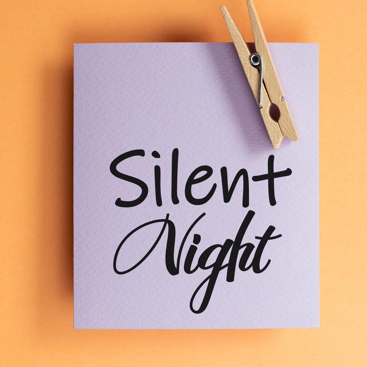 Lavender stamp-and-seal card with black Silent Night script and a wooden clothespin on an orange background.