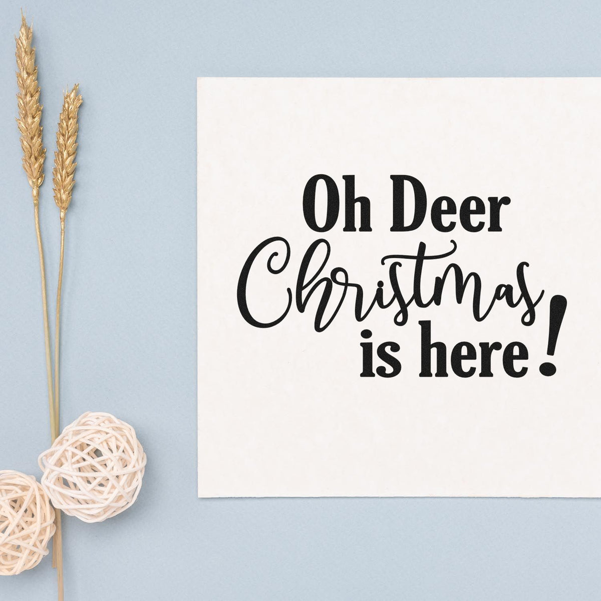 Black Christmas stamp and seal on white card reads Oh Deer Christmas is here! with gold wheat sprigs and white wicker ornaments.