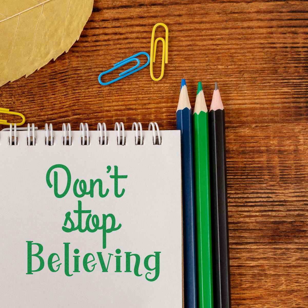 Green motivational stamp and seal on a white spiral notebook, with colored pencils and paper clips on a wood desk.