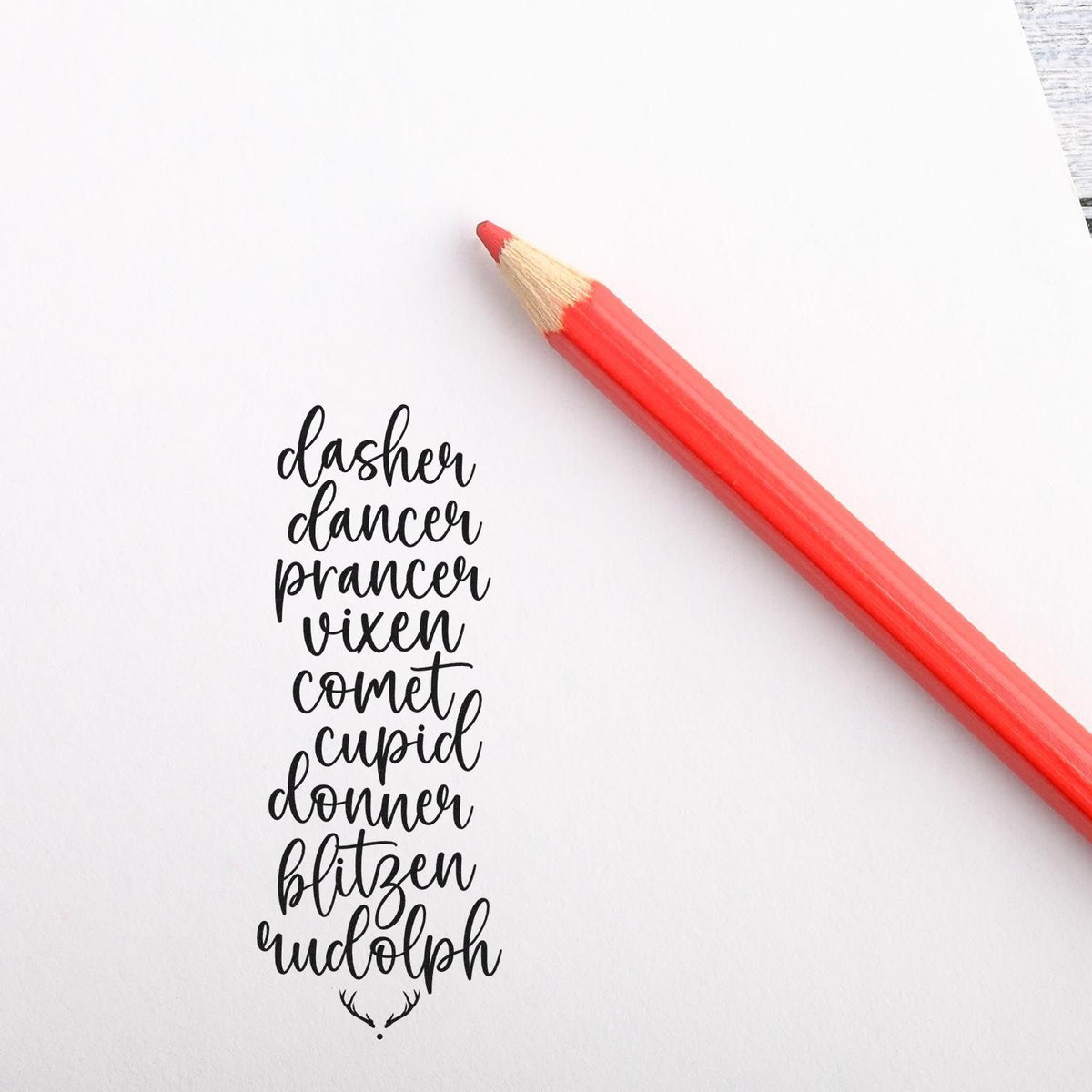 Black Christmas stamp/seal with reindeer names in script, beside a red pencil on white paper.