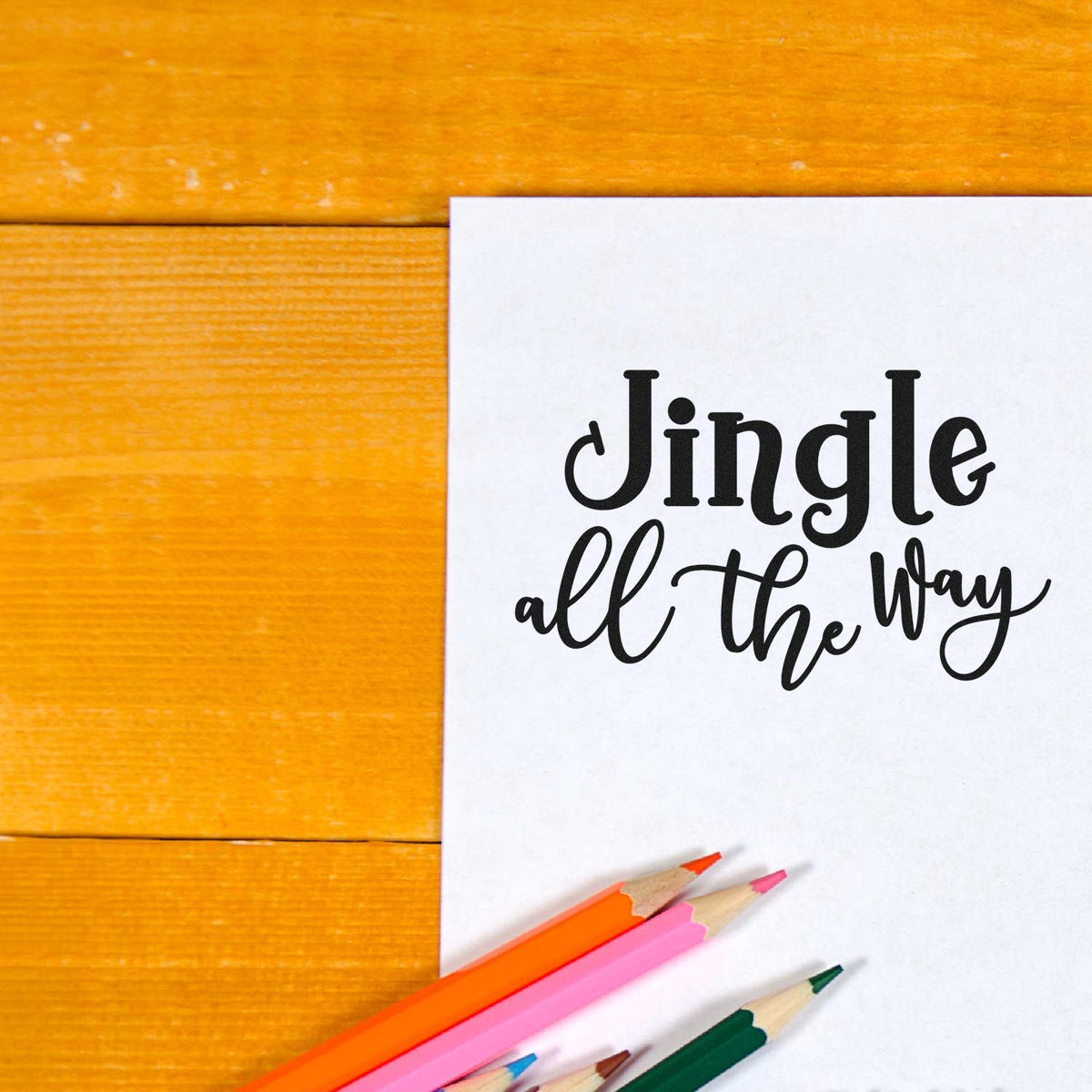Holiday stamp and seal design reading Jingle all the way in black script on white paper, with colorful pencils on orange wood.