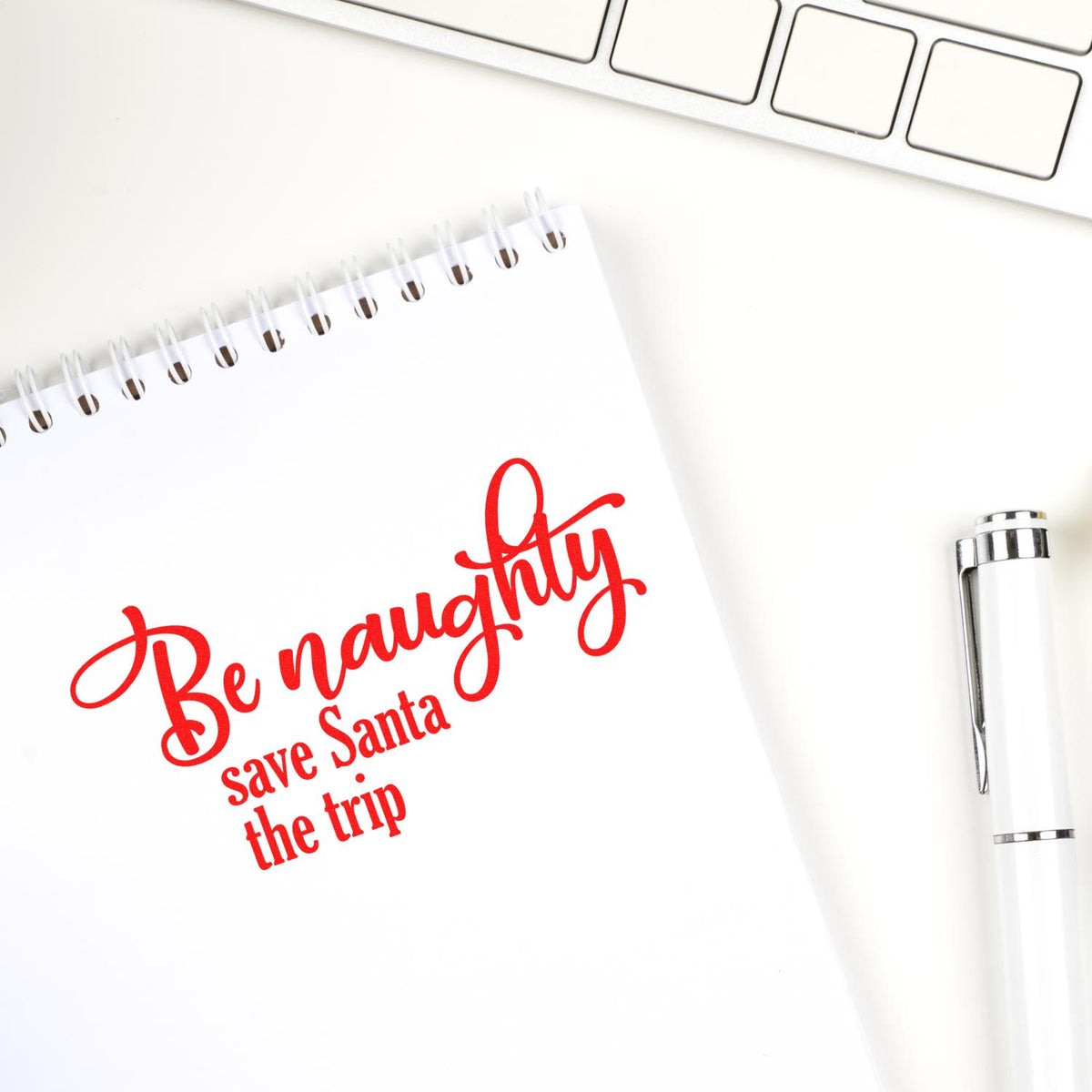White spiral notebook with red holiday stamp reading Be naughty, save Santa the trip beside a white pen and keyboard.