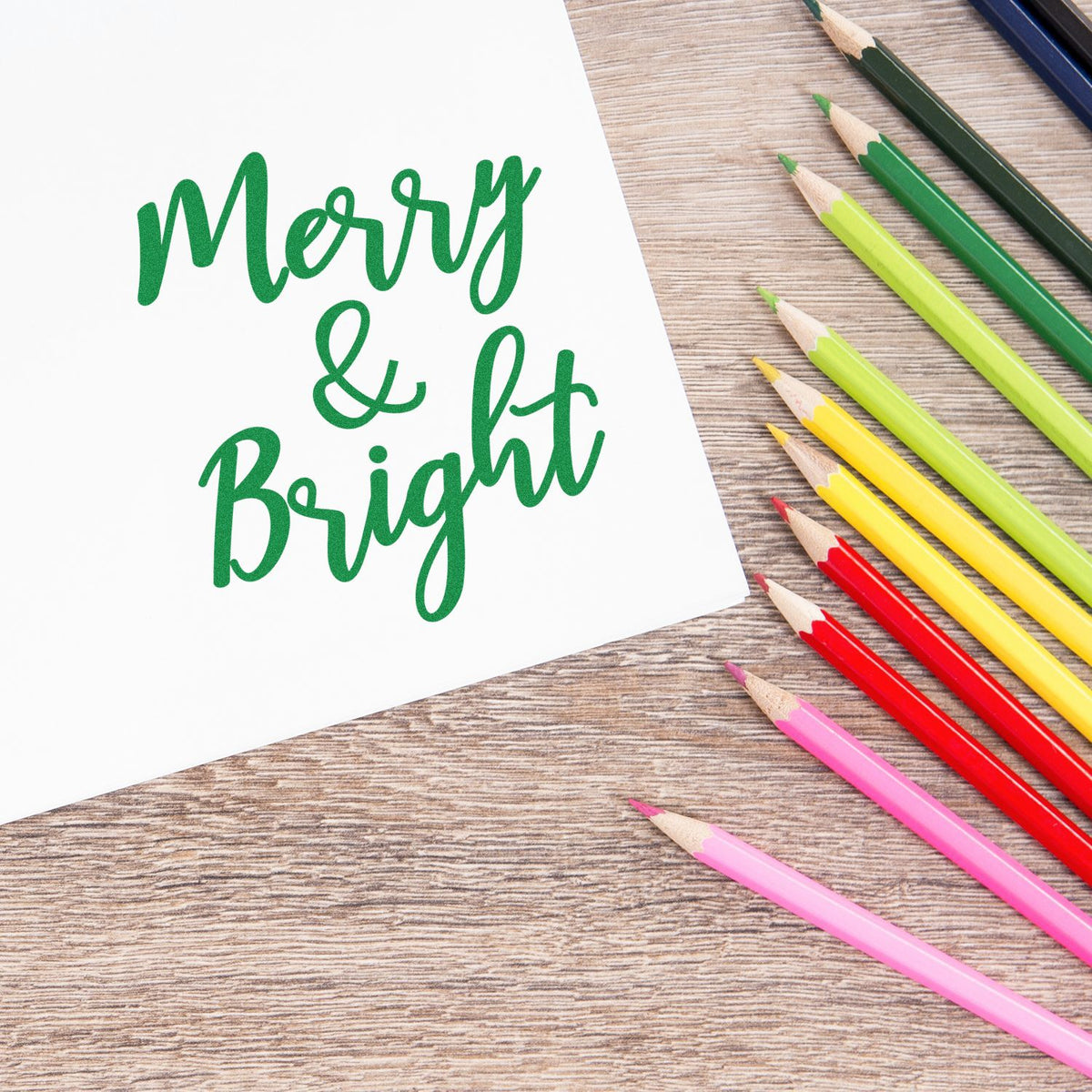 Green Merry & Bright stamp and seal on white paper, surrounded by colorful pencils on a wood table.