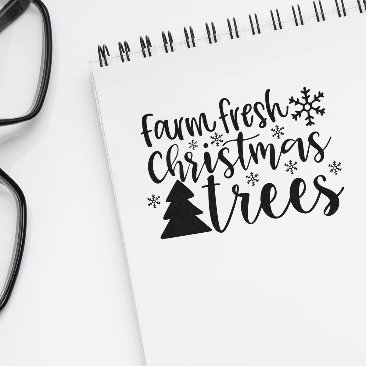 Black farm fresh Christmas trees stamp design with snowflakes and pine tree on a white spiral notebook.