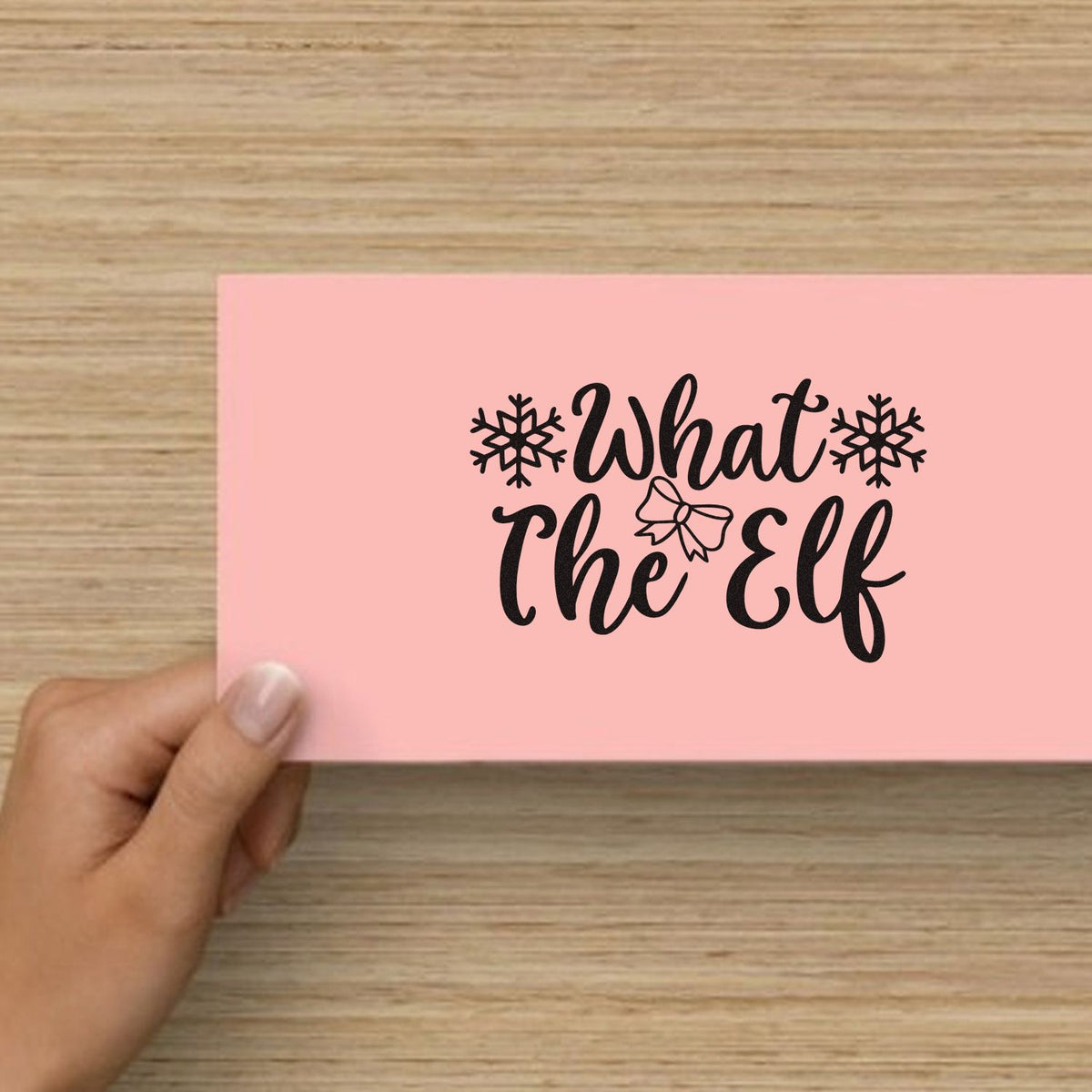 Pink stamp card with black What the Elf holiday seal, snowflakes and bow on a wood background.