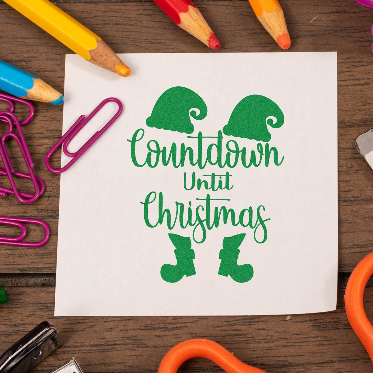 Green Christmas countdown stamp and seal with elf hats and boots on white paper, surrounded by colorful pencils and paper clips.