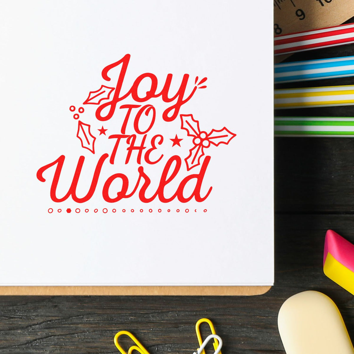 Red holiday stamp and seal design reading Joy to the World with holly accents on white paper, beside colorful pens.