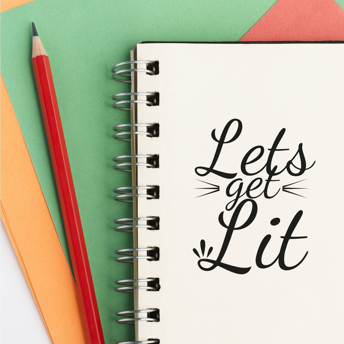 Custom stamp and seal design reading Lets get Lit on a white spiral notebook, with a red pencil and green, orange, and coral paper.