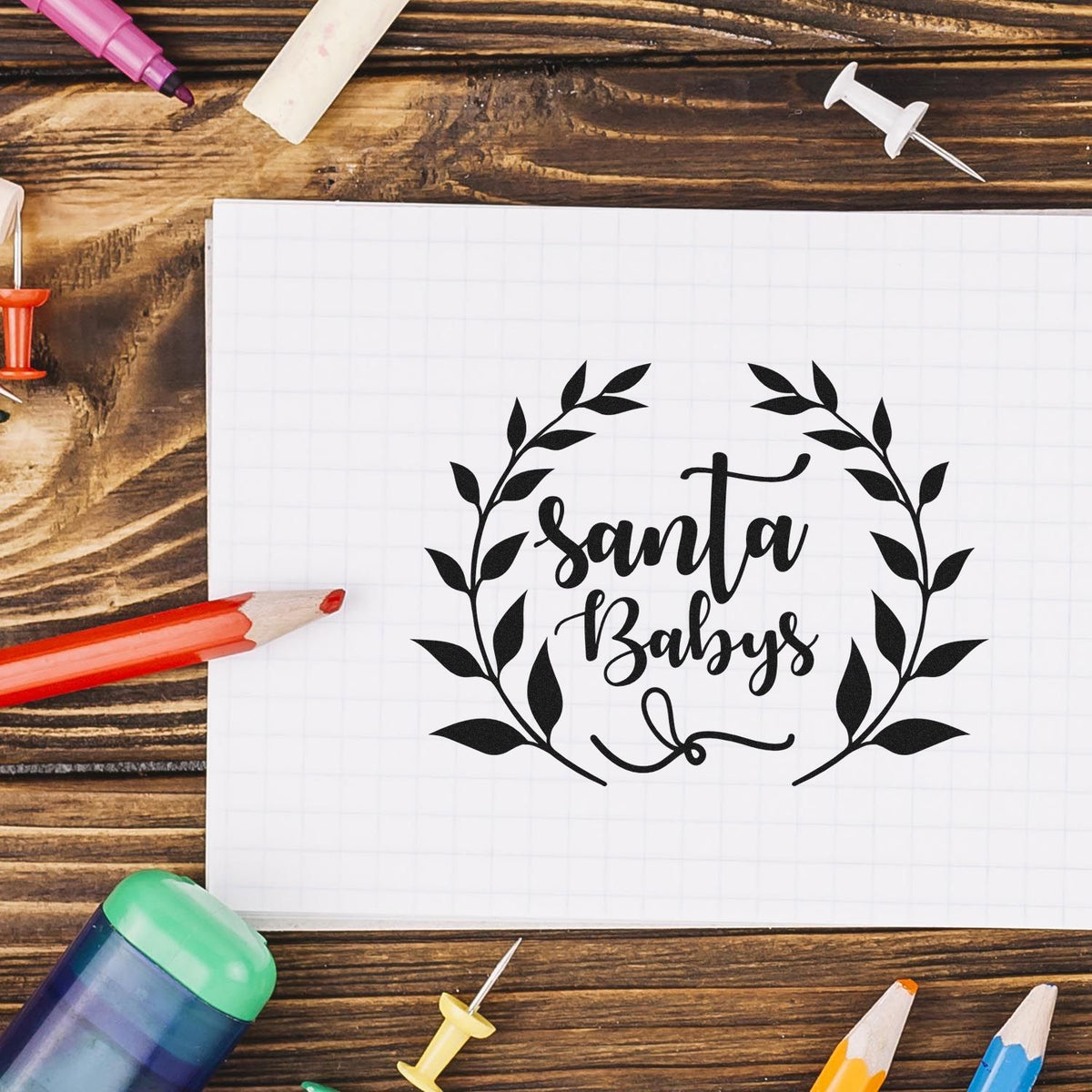 Black Santa Babys stamp and seal design with laurel wreath on white grid paper, surrounded by colorful pencils on rustic wood.