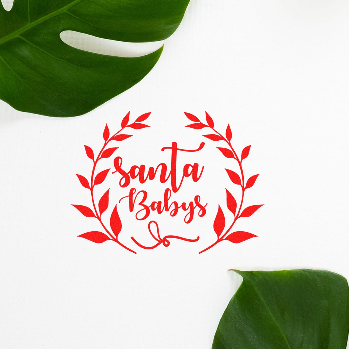 Red Santa Babys stamp and seal in a leafy wreath on white, framed by green leaves.