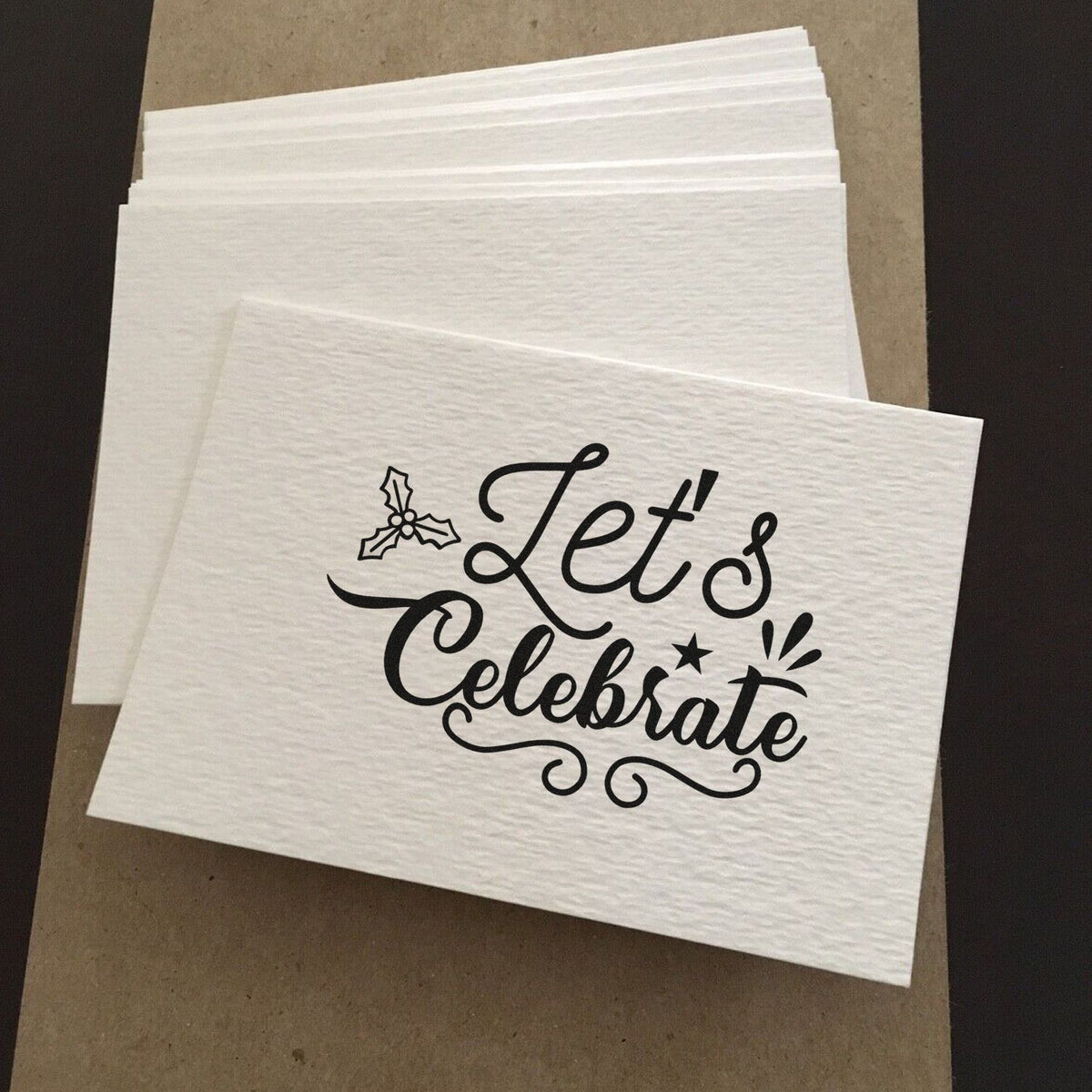 Stacked white textured stamp-and-seal napkins with black Lets Celebrate script, holly accent, and star detail