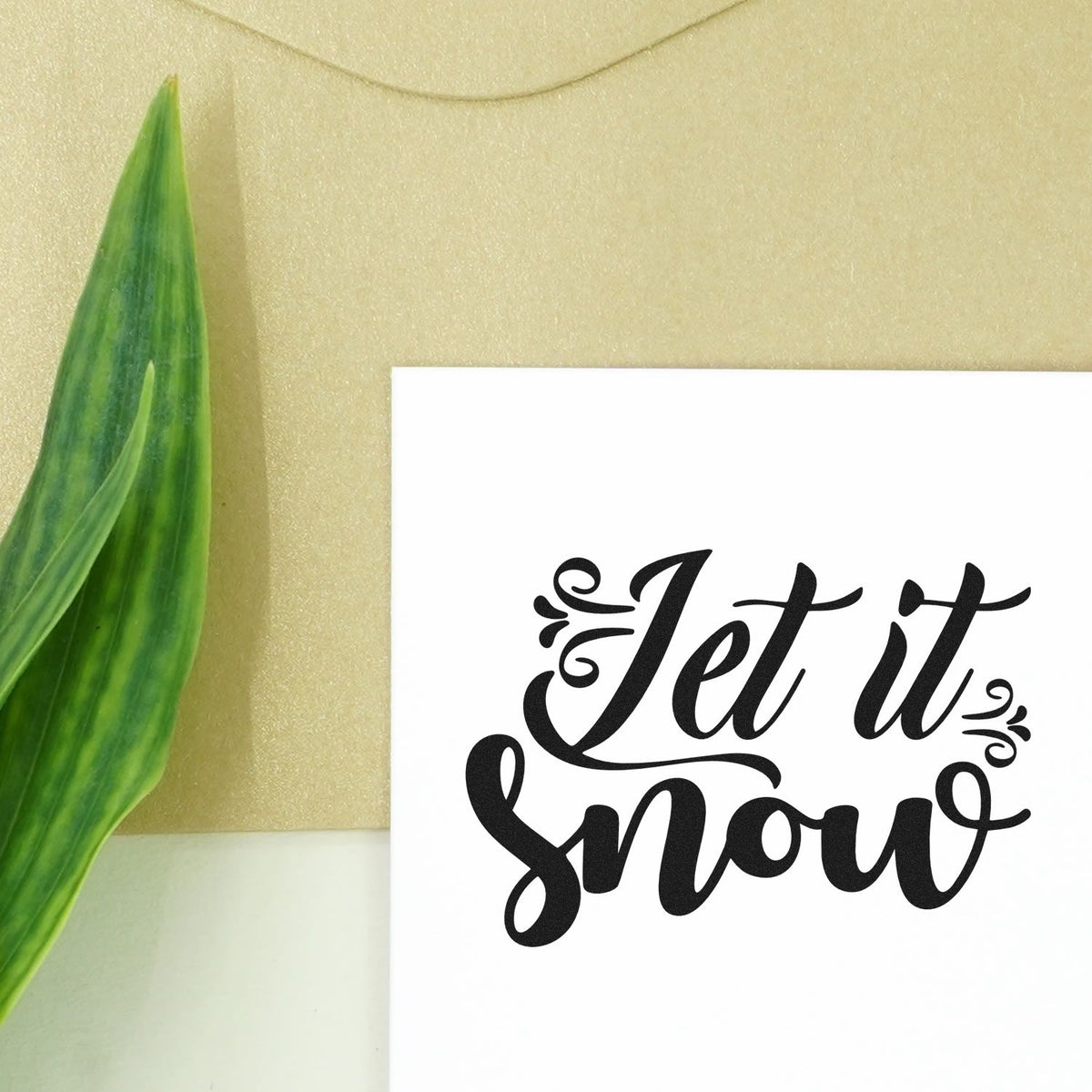Black Let it Snow stamp and seal design on white card, with ornate script on a gold background and green leaf.