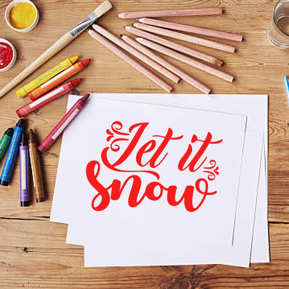 Custom stamp and seal design with red Let it Snow lettering on white paper, surrounded by colored pencils and paint.