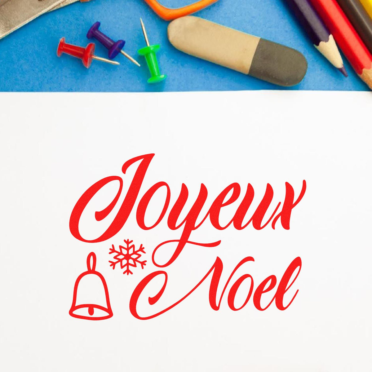 Red Joyeux Noel stamp and seal with bell and snowflake, beside colorful push pins, eraser, and pencils on blue background.