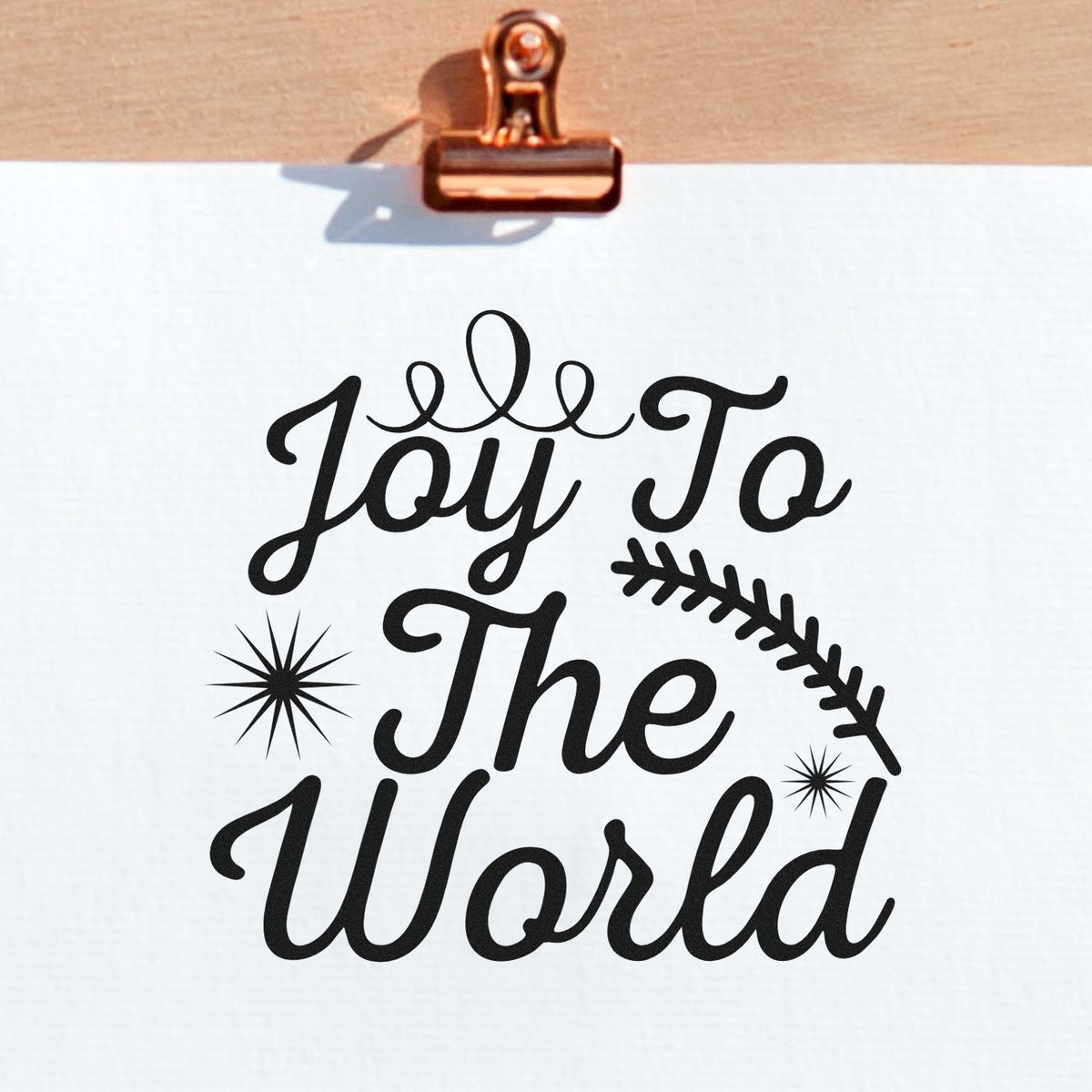 Copper clip holds a white sheet with black Joy To The World custom stamp and seal design, stars and leafy branch.