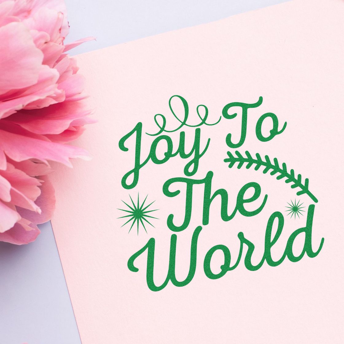 Pastel pink stamp and seal design with green Joy To The World lettering, starbursts, and leafy accents beside pink flowers.