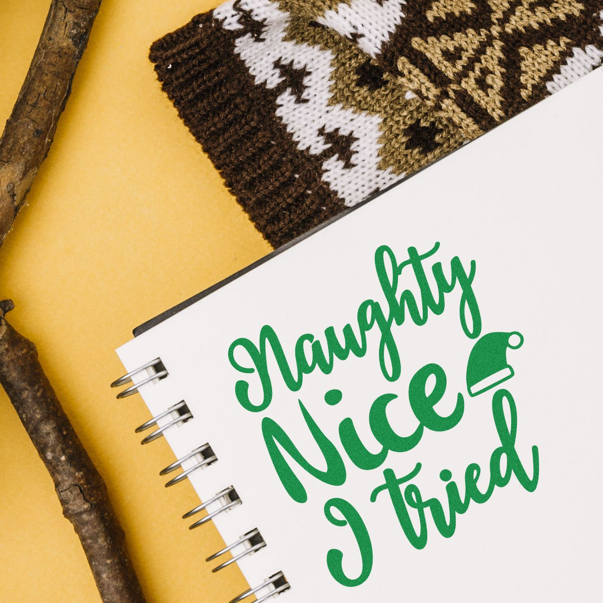 Green Naughty, Nice, Tried stamp seal on a white spiral notebook, with knit fabric and branches on a yellow background.
