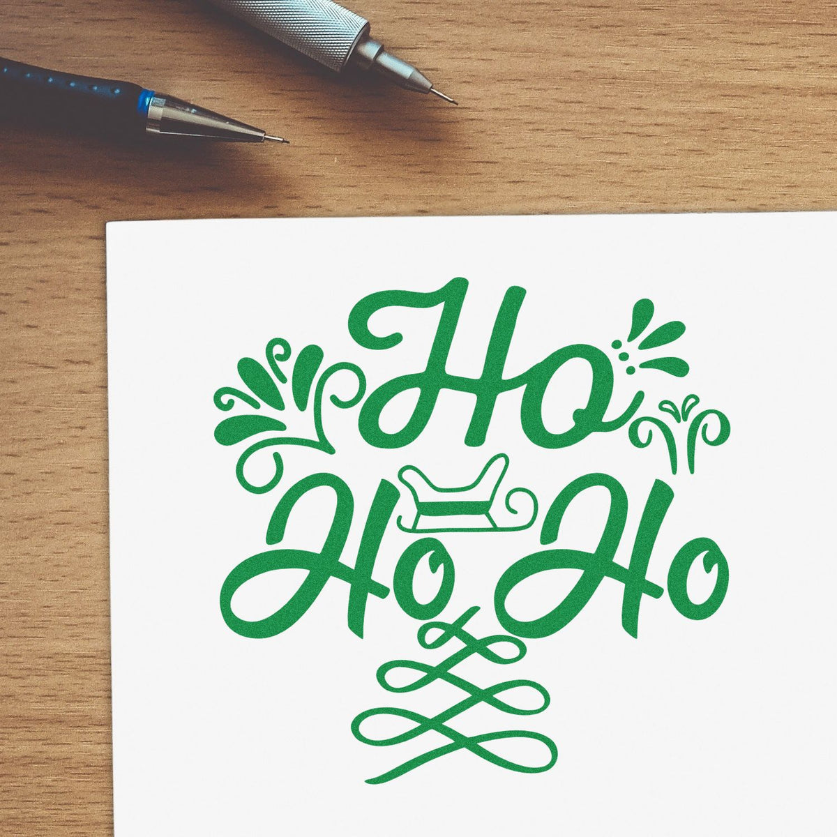 Green decorative Ho Ho Ho stamp and seal design on white paper, with pens on a wooden desk.