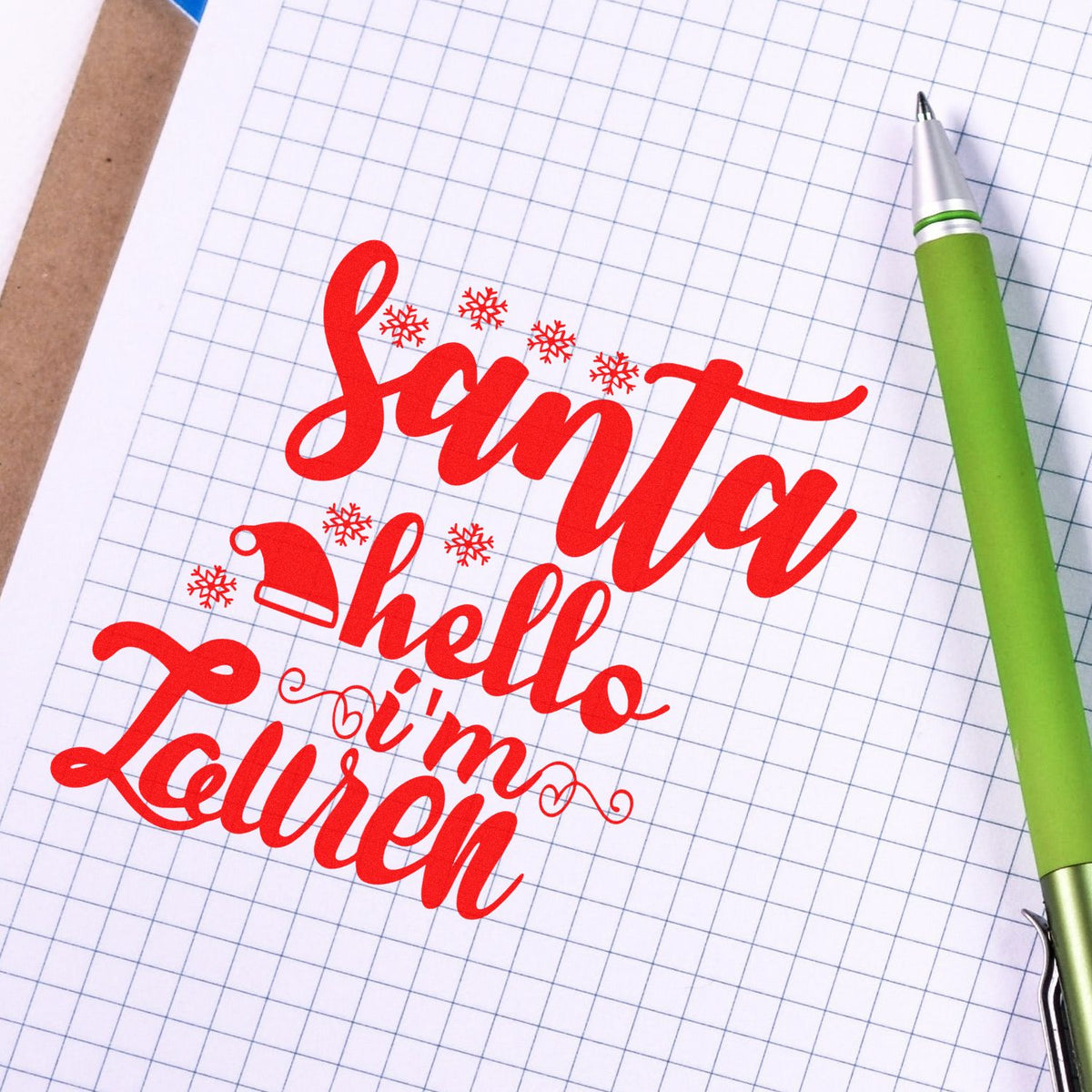 Red custom stamp and seal reading Santa's Helper Lauren with snowflakes and Santa hat on graph paper, beside a green pen.