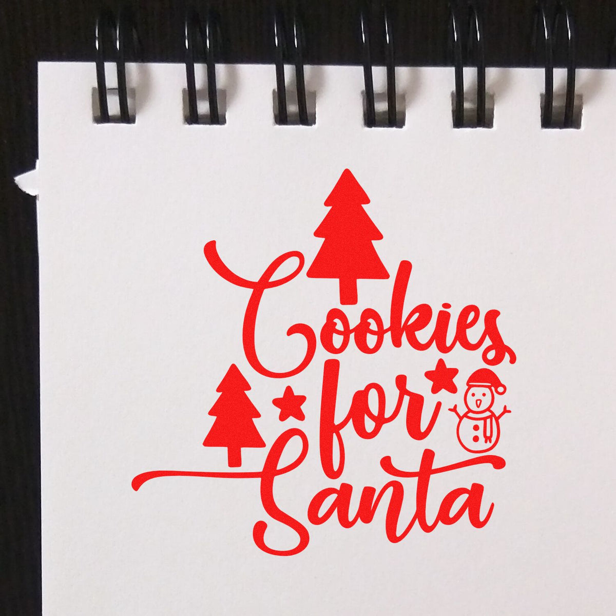 Red holiday stamp seal reading Cookies for Santa with tree, stars, and snowman on a white spiral notebook page.