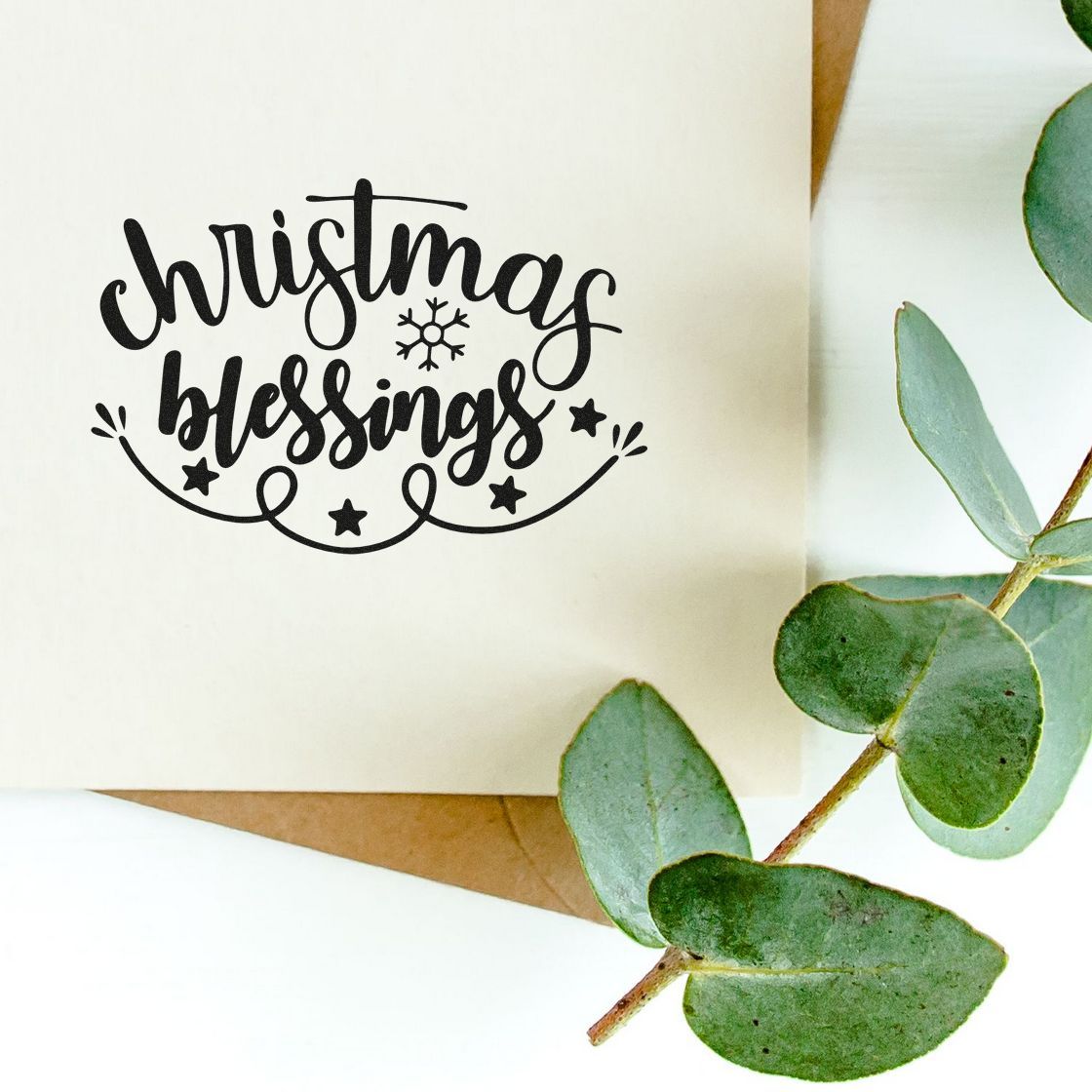 Black Christmas blessings stamp seal on cream paper with snowflake, stars, and green eucalyptus leaves.
