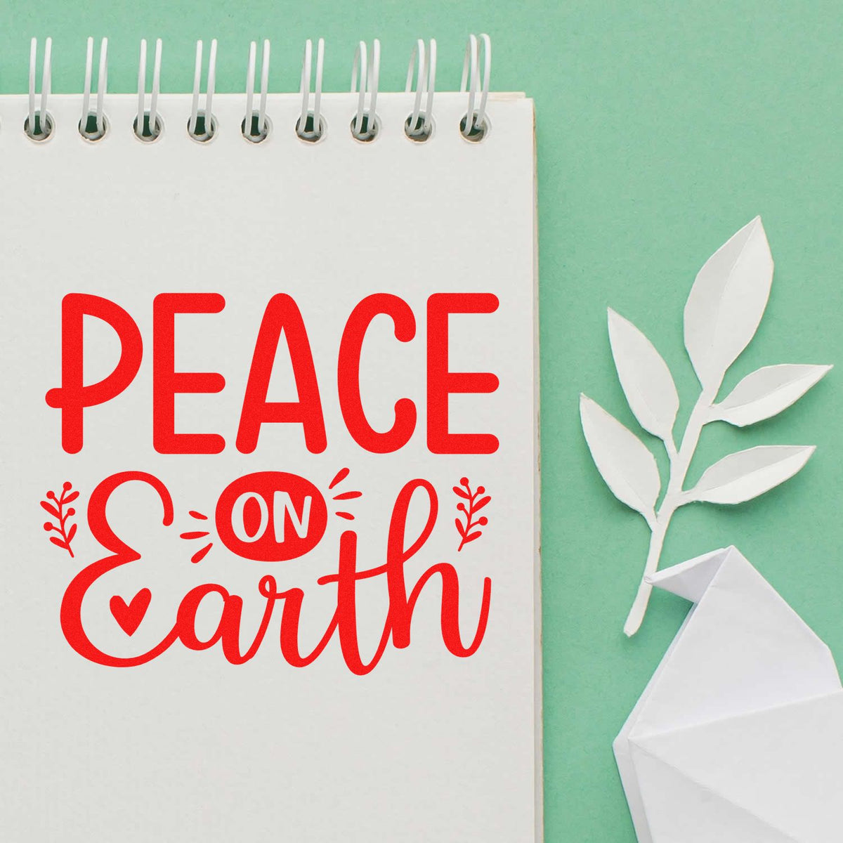 Red "Peace on Earth" stamp and seal design on a white spiral notebook, with a white paper olive branch and origami dove on mint green.