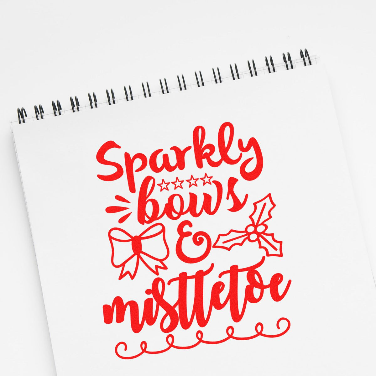 Red Christmas stamp and seal design on a white spiral notebook, reading Sparkly bows & mistletoe with bow and holly accents.