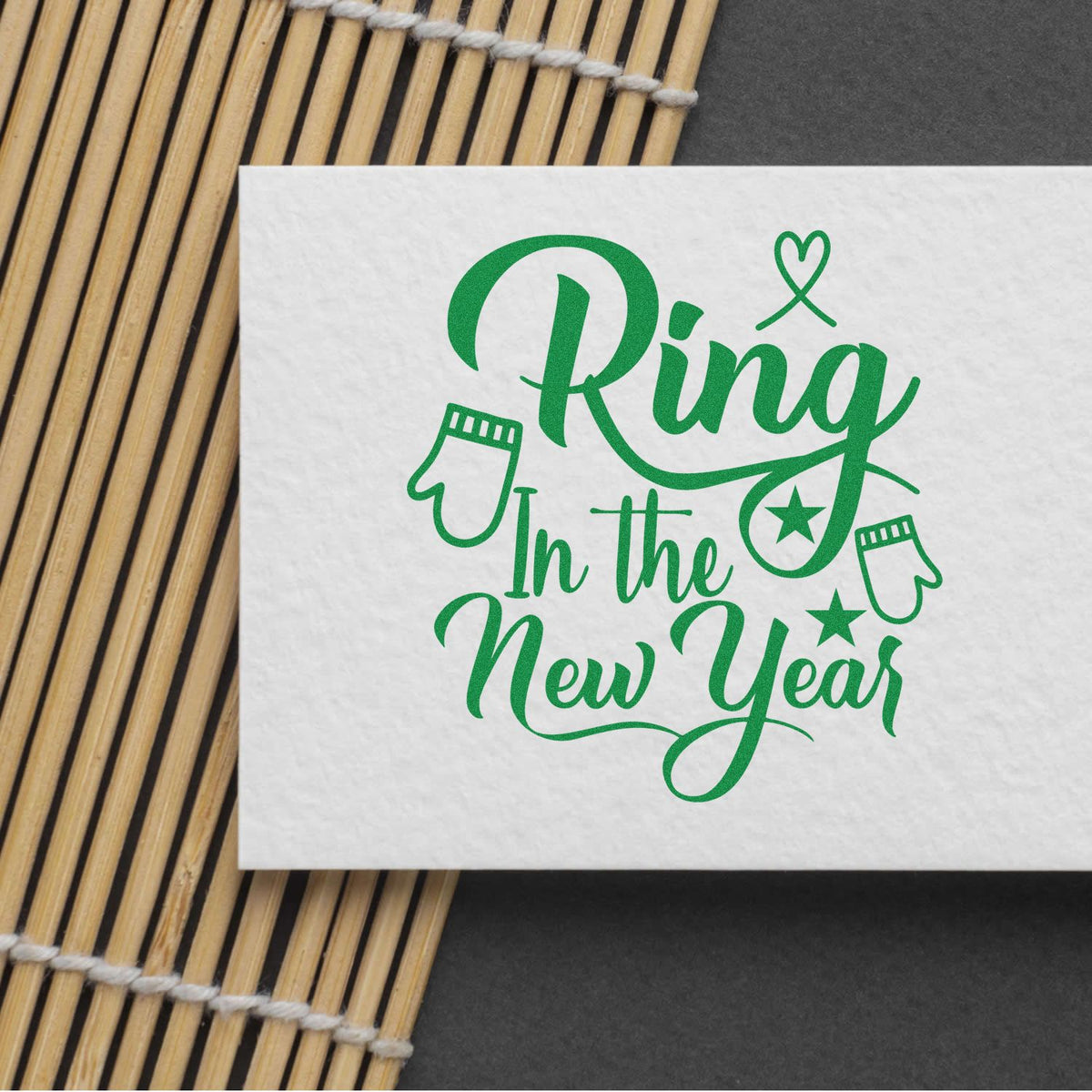 Green Ring in the New Year stamp and seal design with mittens and stars on a white card over a bamboo mat.