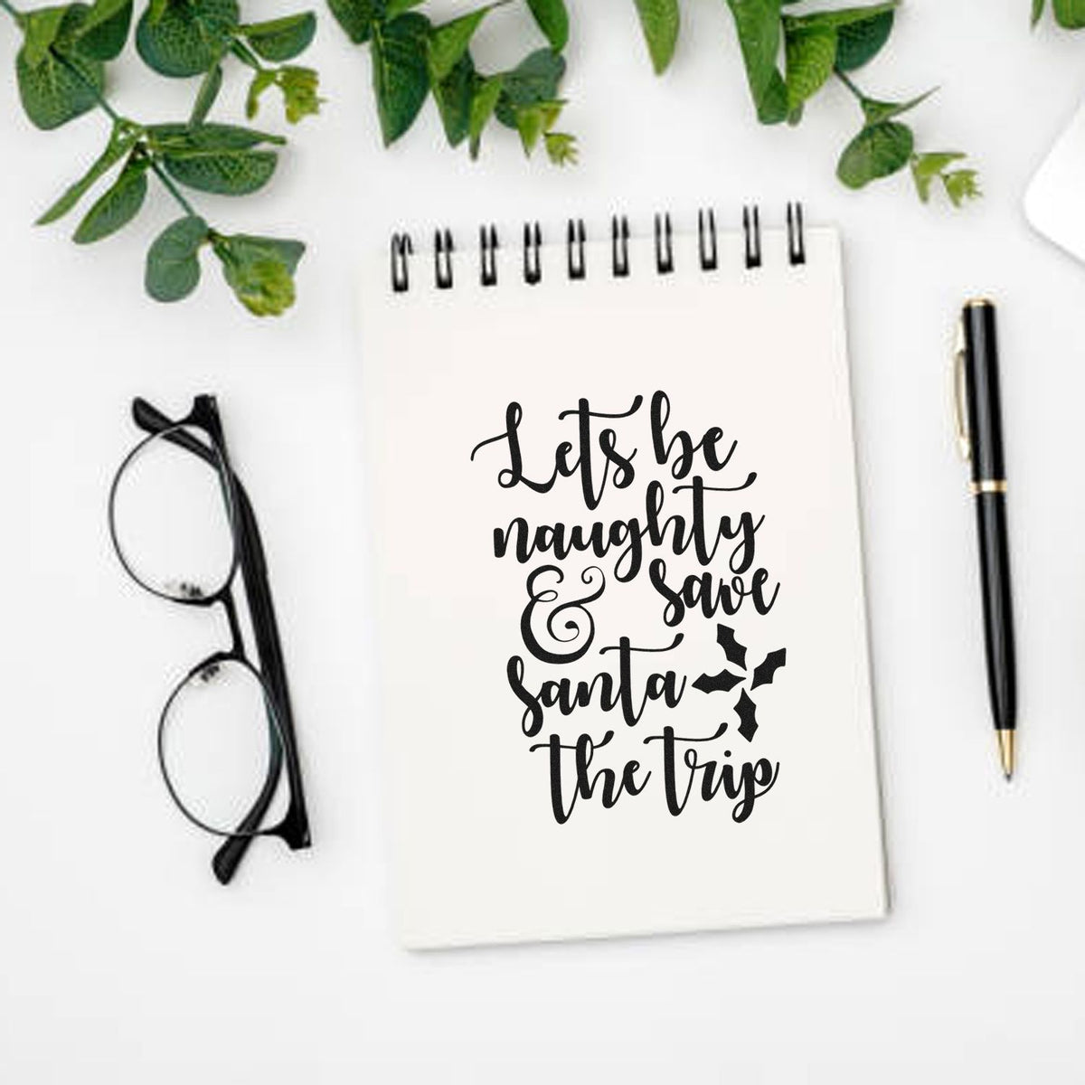Custom stamp and seal design on white spiral notebook, black script reading Lets be naughty & save Santa the trip, with pen and glasses.