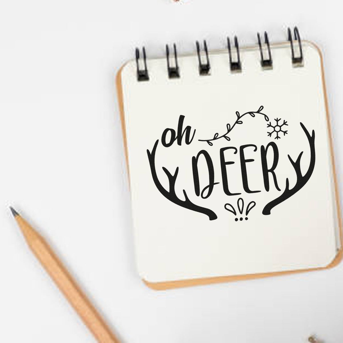 Black oh DEER stamp design with antlers and snowflake on a cream notepad, beside a wooden pencil.