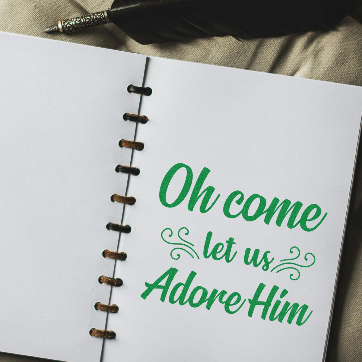 Green script stamp and seal design on a white spiral-bound page reading Oh come let us Adore Him beside a quill pen.