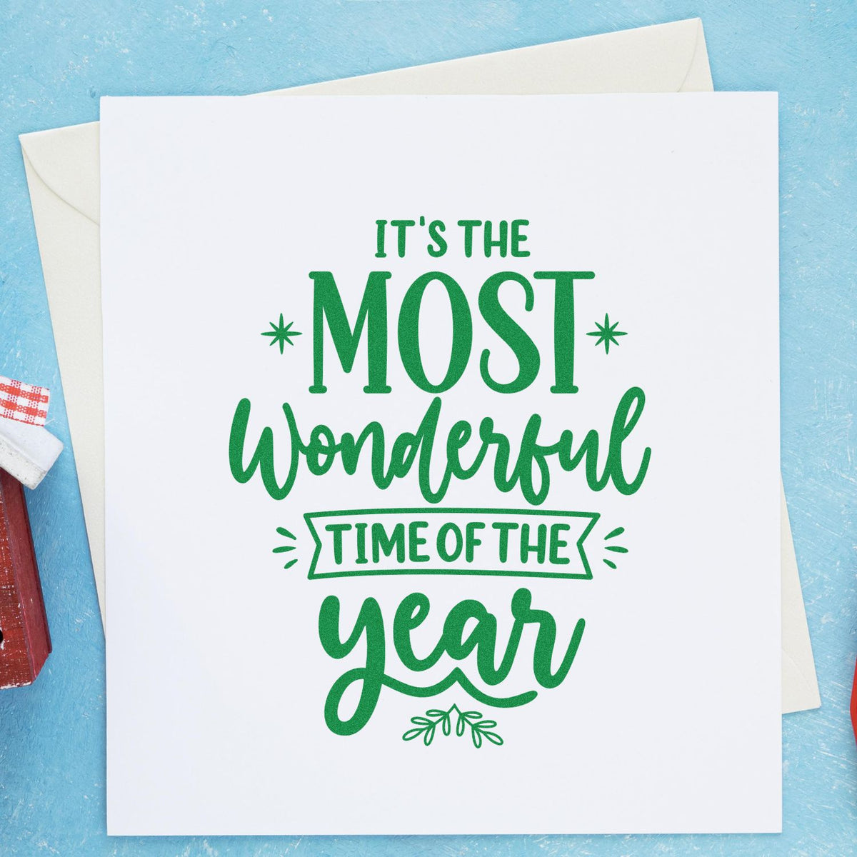 Green holiday stamp and seal design on white card reading Its the most wonderful time of the year, with envelopes on blue background.