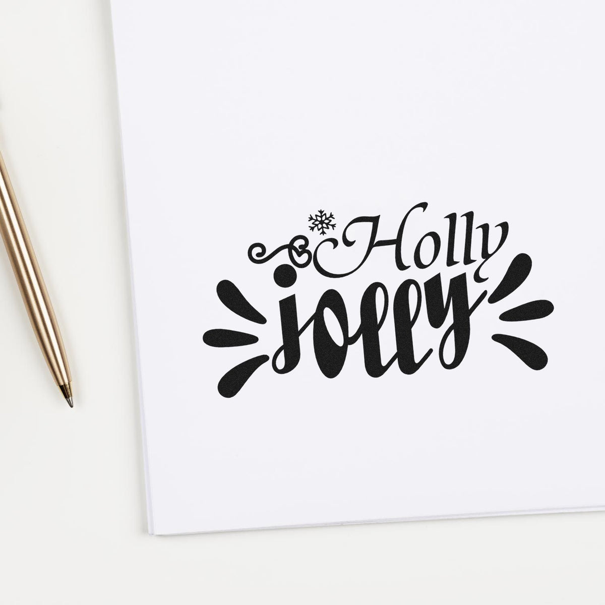 Black Holly Jolly rubber stamp design on white paper with snowflake accents, beside a gold pen.