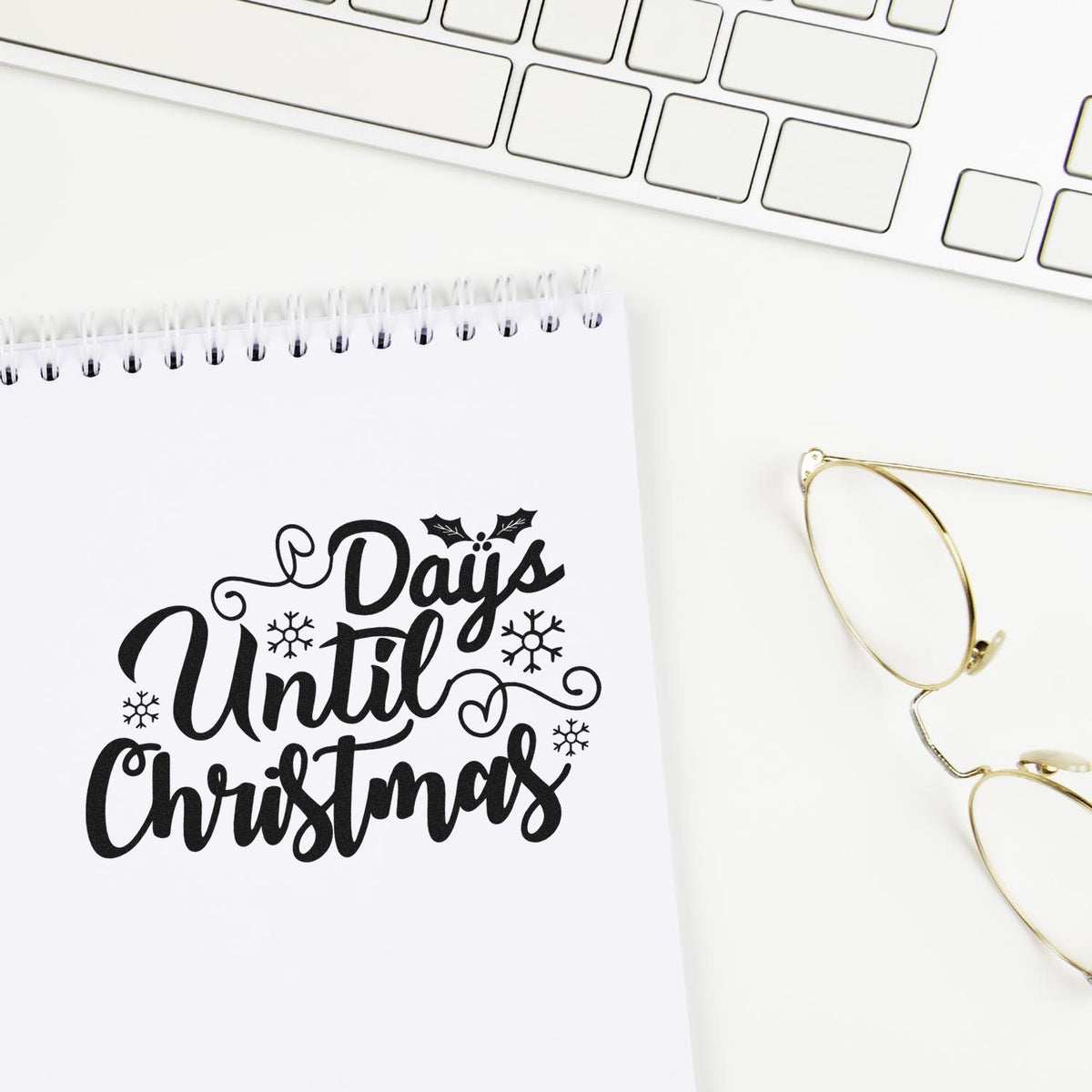 Black holiday stamp and seal design reading Days Until Christmas with snowflakes on a white notebook beside a keyboard and gold glasses.