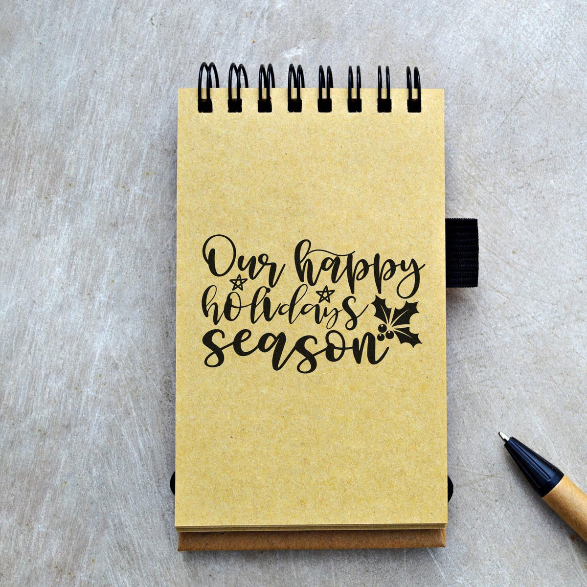 Yellow spiral notepad featuring a black holiday stamp reading Our happy holidays season with holly; stamp and seal product.