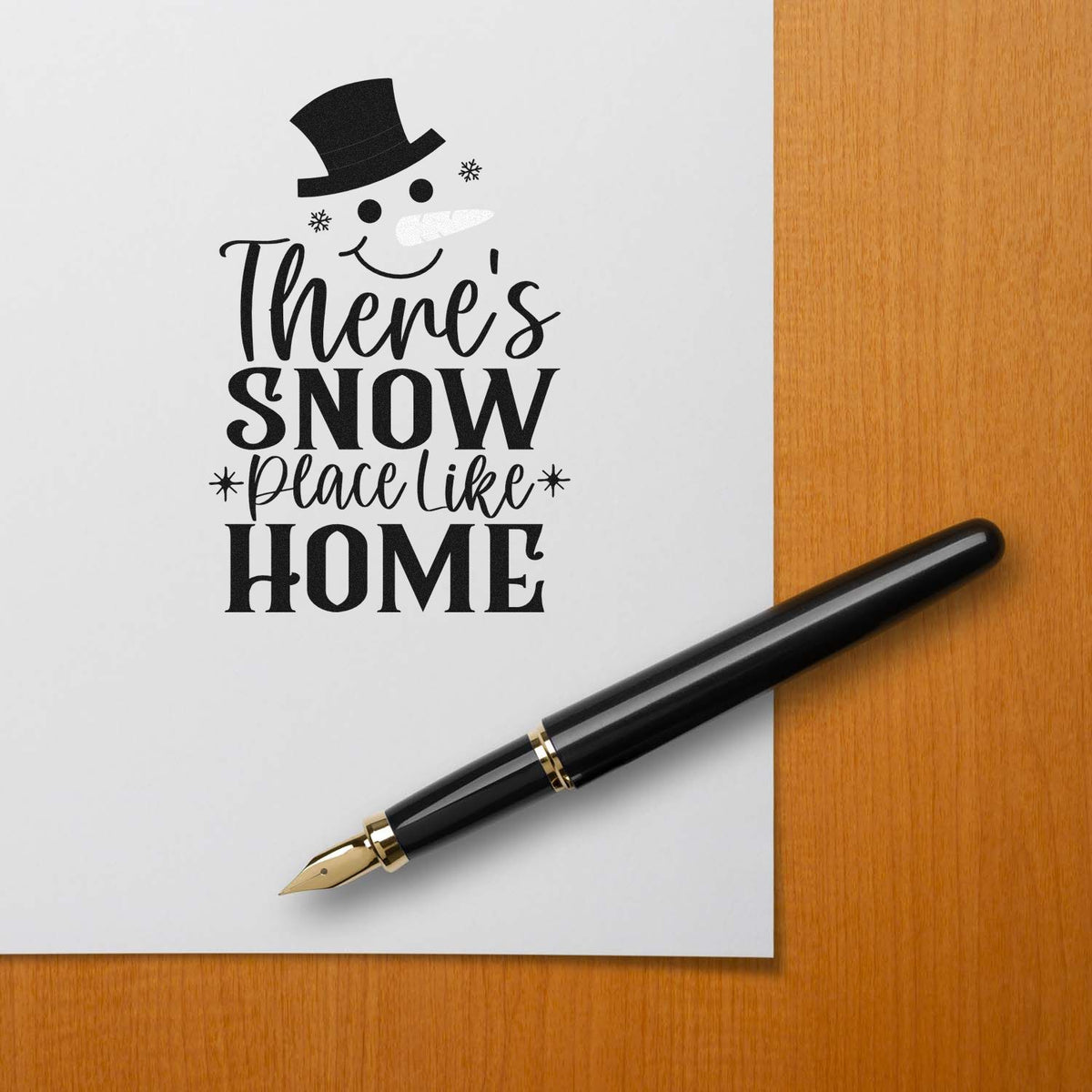 Black stamp and seal design with snowman hat and Theres snow place like home beside a black fountain pen on white paper.