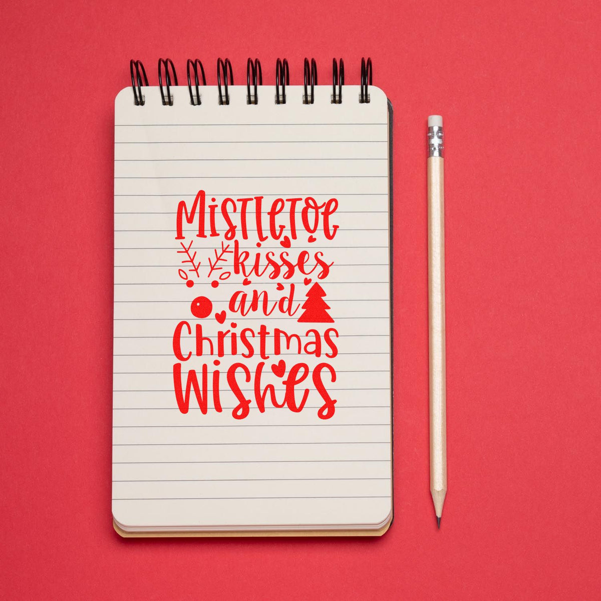 Red Christmas stamp on a lined spiral notebook with pencil, reading Mistletoe kisses and Christmas wishes.