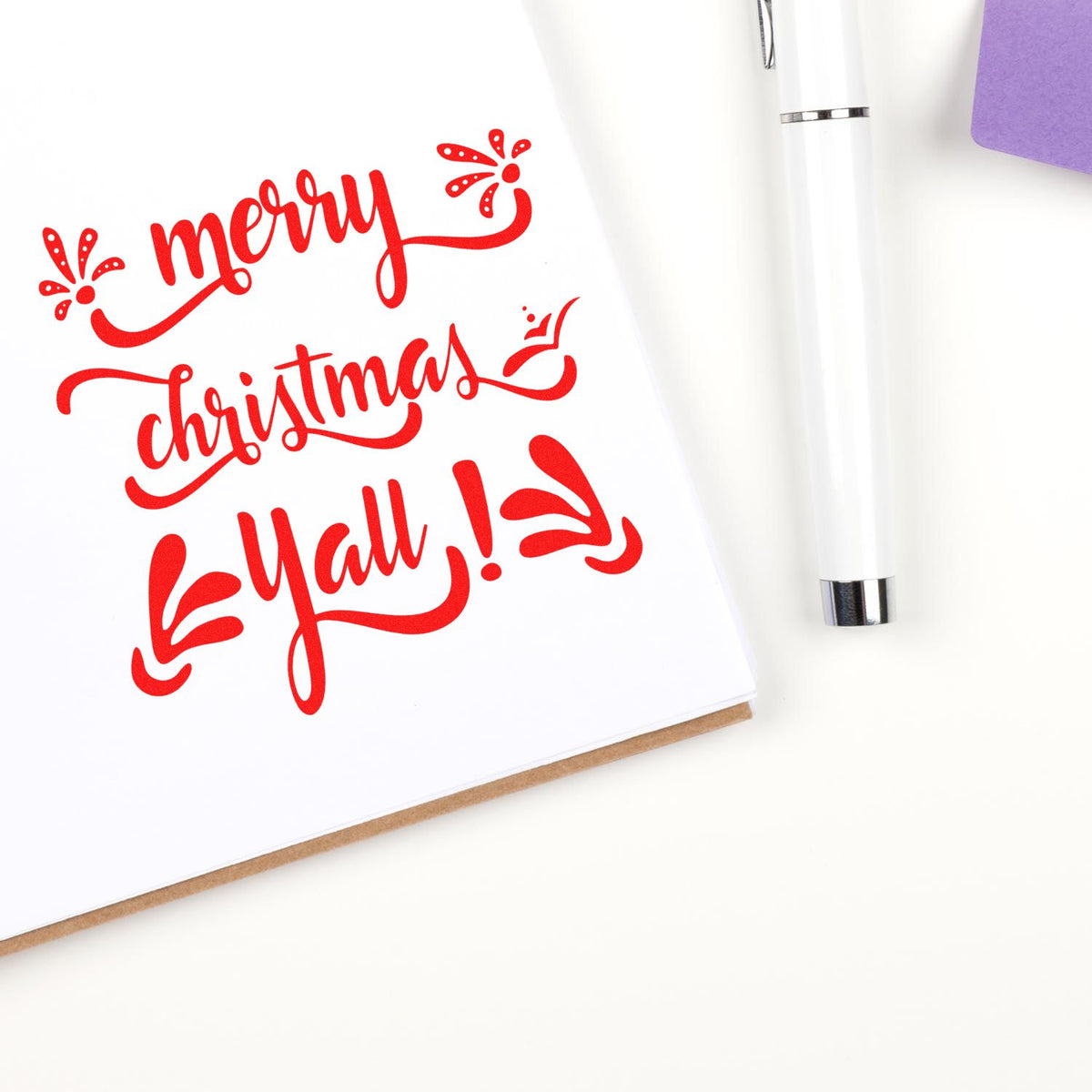 Red Merry Christmas stamp design with decorative swirls, beside a white seal pen and purple card.