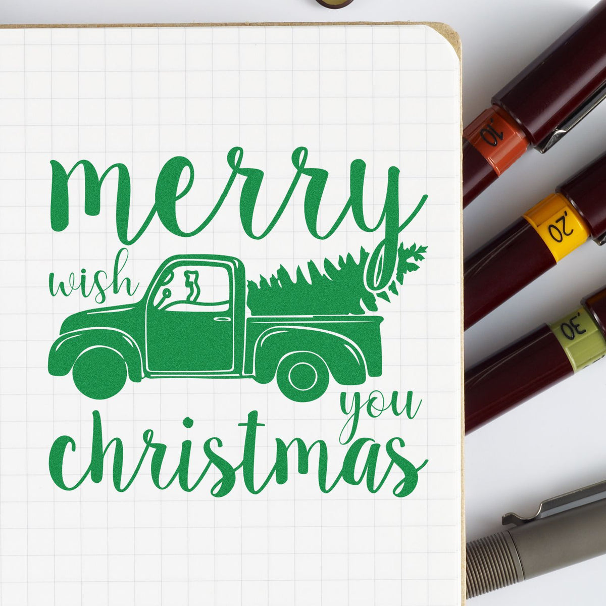 Green Christmas stamp design with pickup truck and tree on grid notebook, beside maroon, yellow, and olive stamp and seal markers.