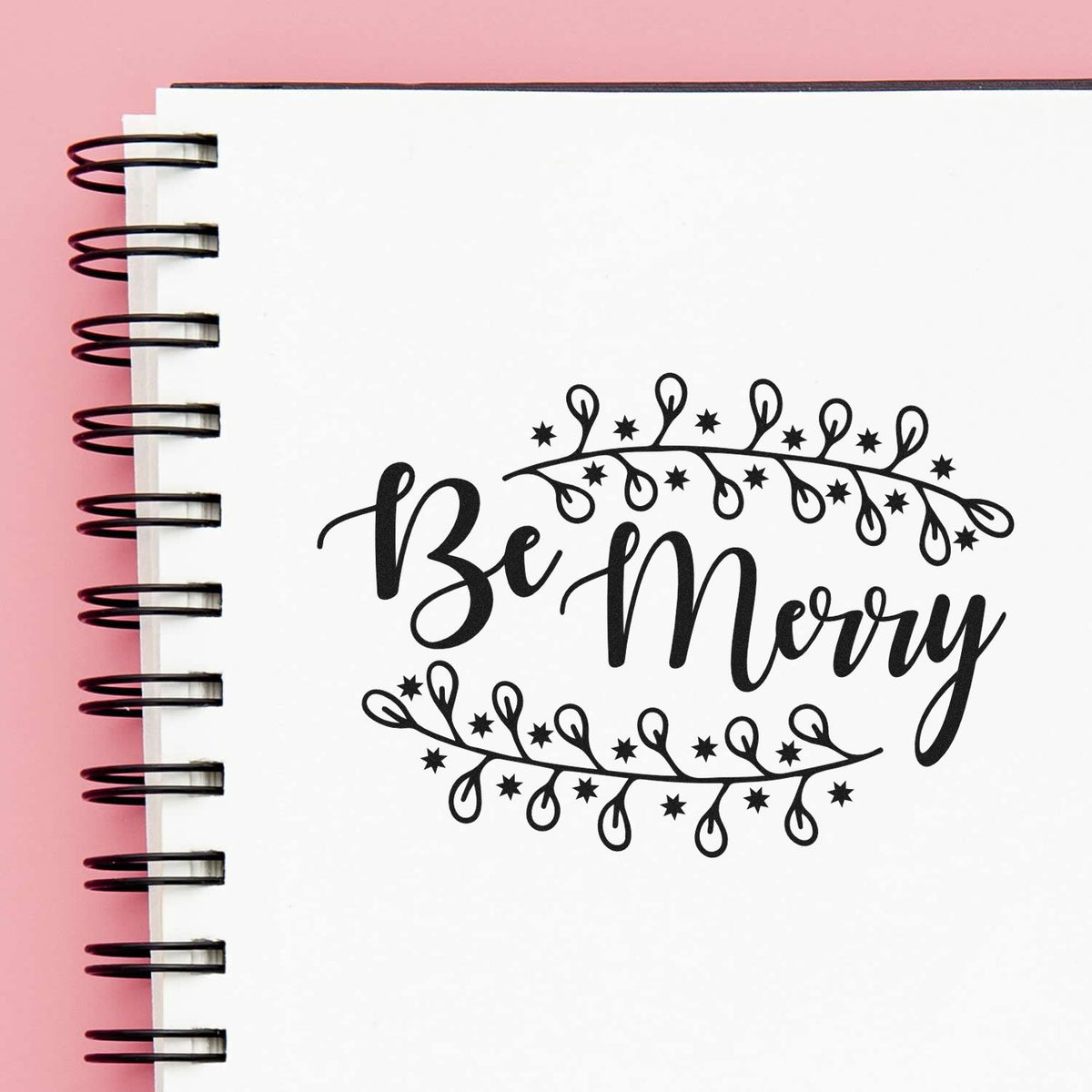 Black Be Merry holiday stamp design with stars and leafy wreath on white notebook, against a pink background.
