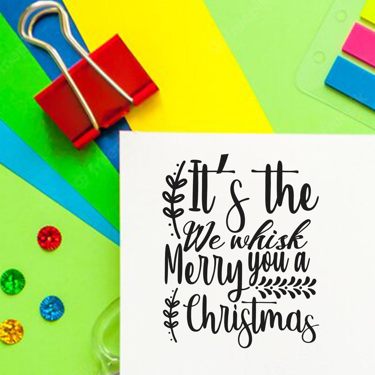 Colorful stamp and seal products with red binder clip and black Christmas greeting on white card against neon paper.