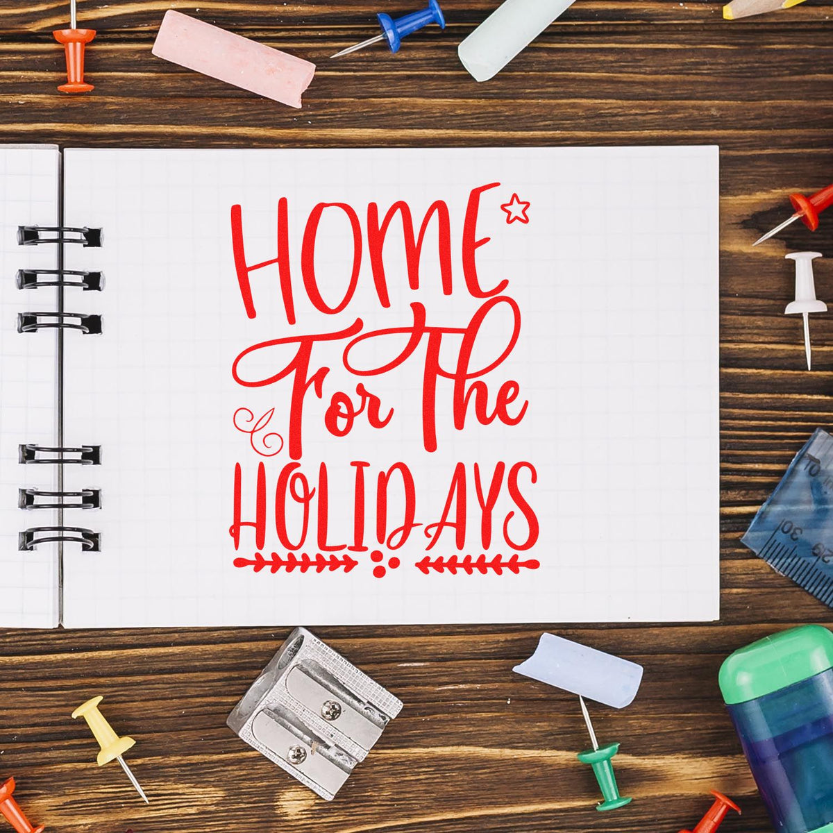 Red Home for the Holidays stamp and seal design on open notebook, with colorful push pins and desk supplies on wood.