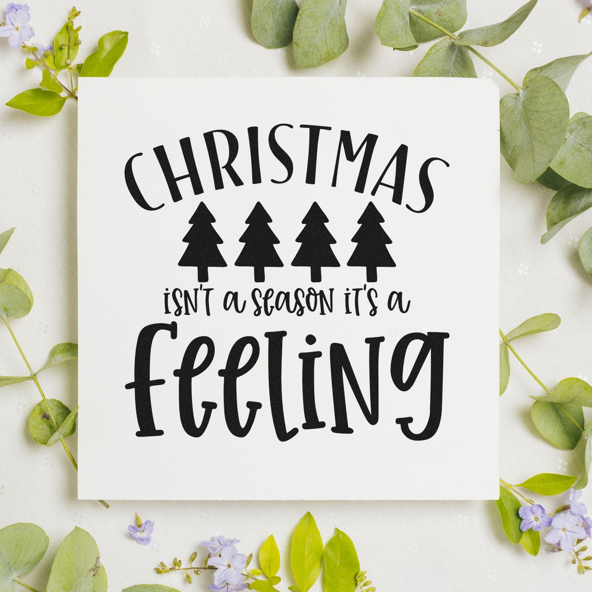 Black Christmas stamp seal with four pine trees and isnt a season its a feeling on white, framed by green leaves and lilac flowers
