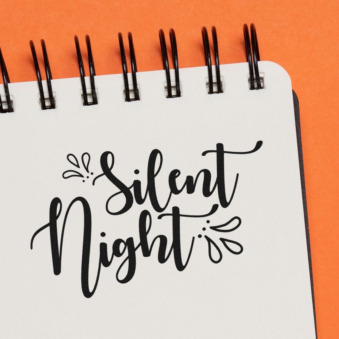 Black Silent Night stamp/seal design on a white spiral notebook page, with decorative swirls on an orange background.