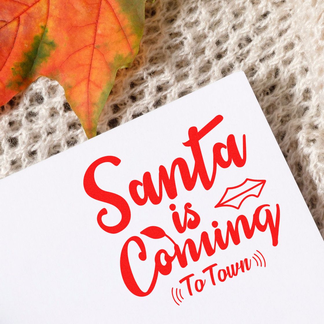 Red custom stamp and seal reading Santa is Coming To Town on white paper, with autumn leaf and beige knit background.