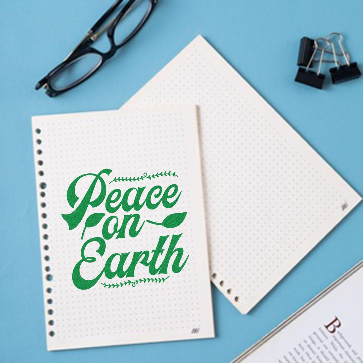 Green Peace on Earth decorative stamp on white dotted pages, with black glasses and binder clips on a blue background.