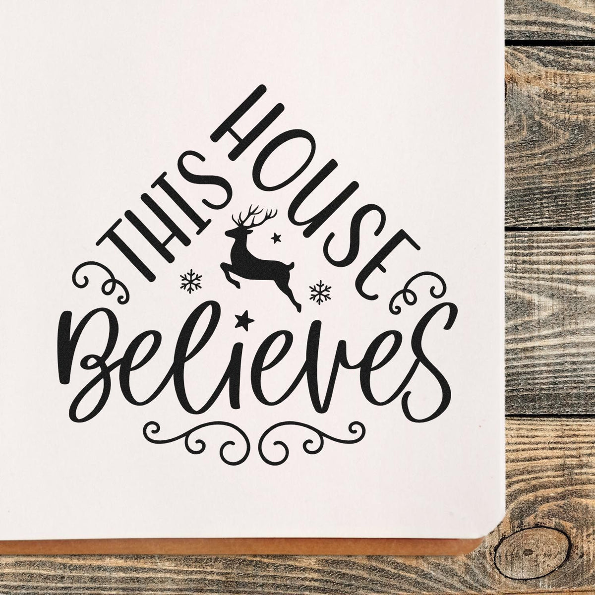 Black holiday stamp/seal with This House Believes, deer, snowflakes, and swirls on white paper over rustic wood.